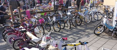 Eine große Anzahl von Kinderfahrrädern ist auf einer gepflasterten Straße geparkt, vor einem Markt mit verschiedenen ausgestellten Artikeln, umgeben von Menschen.