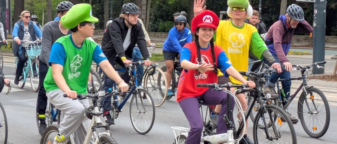 Eine Gruppe von Radfahrern in grünen Mario-Kart-Kostümen fährt an einem sonnigen Tag ihre Fahrräder. Bäume und Straßenlampen säumen die Straße.