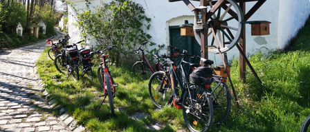 Eine Reihe von Fahrrädern ist vor einem rustikalen Gebäude mit einem Brunnen und einem Rad unter einem hölzernen Vordach geparkt.