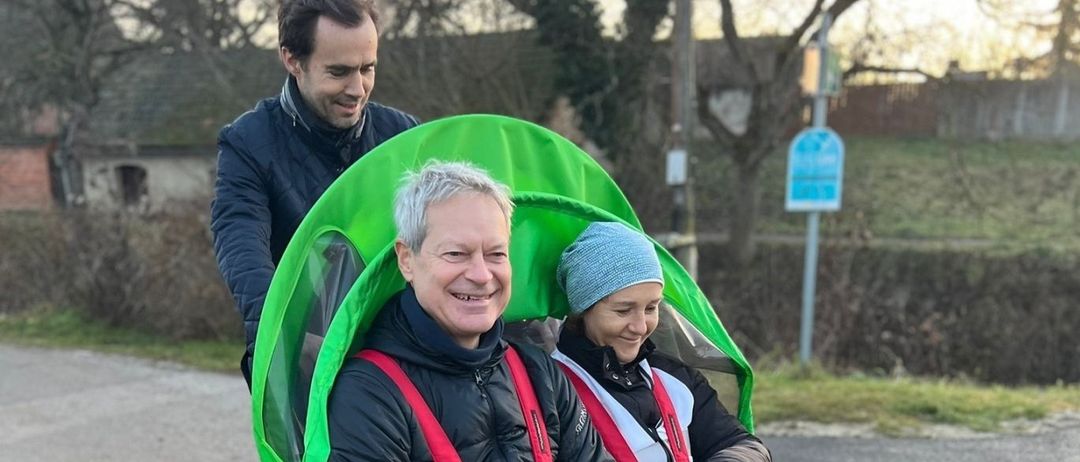 Ein älterer Mann sitzt in einem Rollstuhl mit grüner Abdeckung und lächelt. Eine Frau neben ihm trägt eine Mütze und Handschuhe. Sie fahren auf einer Straße, mit einem Mann hinter ihnen. Bäume und ein Haus sind im Hintergrund.