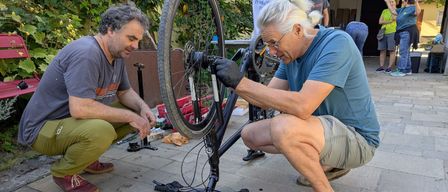 Zwei Männer reparieren einen Fahrradreifen auf einem gepflasterten Gehweg. Ein Mann hält den Reifen, während der andere ihn justiert. In der Nähe steht eine Person, und im Hintergrund befinden sich ein Banner und Pflanzen.