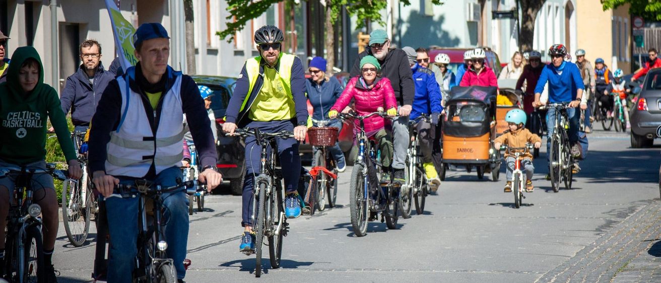 Eine Gruppe von Radfahrern fährt eine Stadtstraße entlang, einige mit Helmen und andere mit Kindern auf Fahrrädern. Bäume und Gebäude säumen die Straße, und in der Nähe befindet sich ein Kinderwagen mit einem Kind.