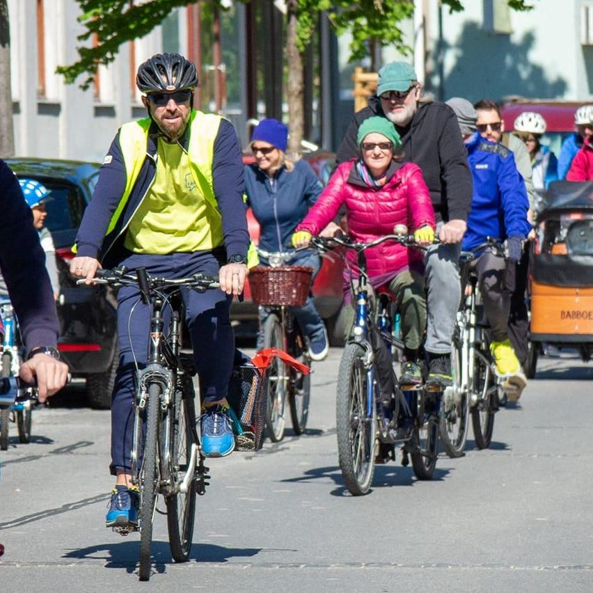 Eine Gruppe von Radfahrern fährt eine Stadtstraße entlang, einige mit Helmen und andere mit Kindern auf Fahrrädern. Bäume und Gebäude säumen die Straße, und in der Nähe befindet sich ein Kinderwagen mit einem Kind.