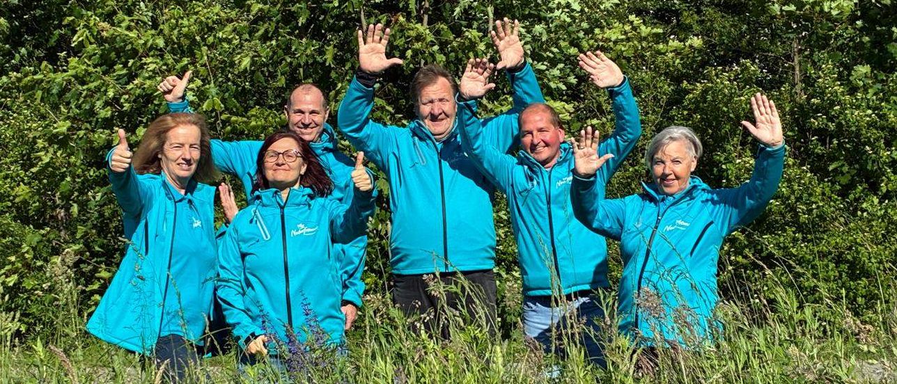 Eine Gruppe von sechs Personen in blauen Jacken steht in einem Grasfeld und lächelt. Der Hintergrund ist mit dichten grünen Bäumen gefüllt.