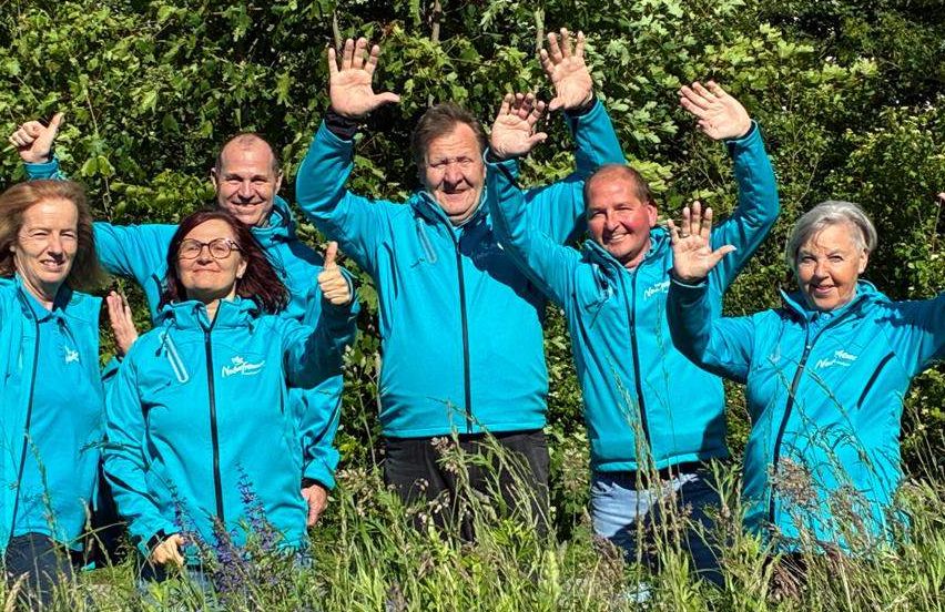 Eine Gruppe von sechs Personen in blauen Jacken steht in einem Grasfeld und lächelt. Der Hintergrund ist mit dichten grünen Bäumen gefüllt.