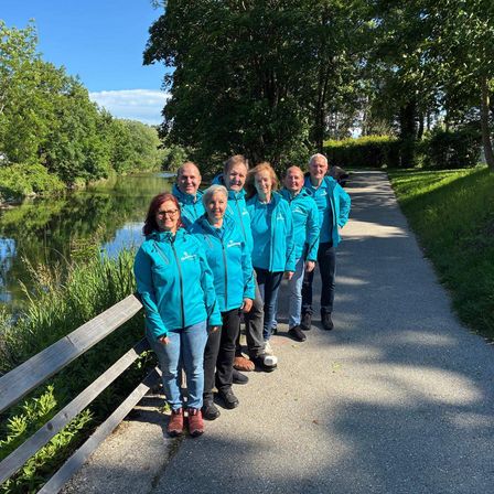Eine Gruppe von sieben Personen, alle in blauen Jacken, posiert für ein Foto am See an einem sonnigen Tag.