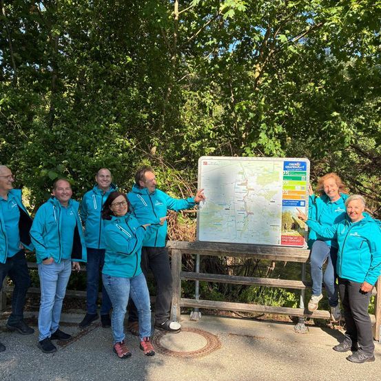 Eine Gruppe von Menschen in passenden blauen Jacken posiert für ein Foto und zeigt auf eine Karte. Die Karte ist auf einem Holzgestell montiert. Hinter ihnen befinden sich Bäume und Grünpflanzen.