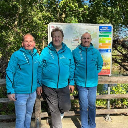 Drei Männer in blauen Jacken posieren für ein Foto vor einer Karte und einem Holzzaun in der Nähe eines Flusses.