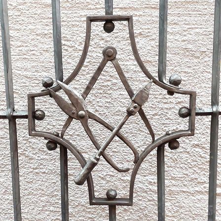 Ein Nahaufnahme eines metallischen Gitterdesigns mit einem zentralen Emblem aus gekreuzten Scheren und einem Hammer, vor einem strukturierten Hintergrund.