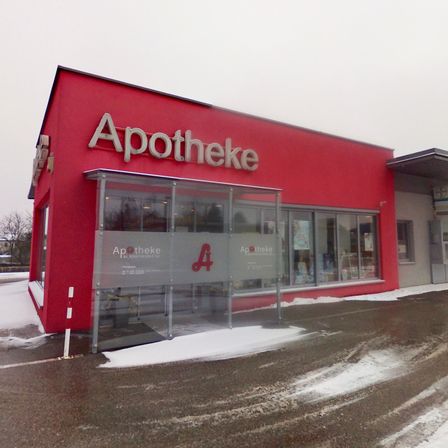 Eine Apotheke mit rotem Äußeren und Glaseingang, ausgestattet mit einem Glasvordach. Im Inneren sind Regale mit verschiedenen Artikeln zu sehen. Das Gebäude ist schneebedeckt.