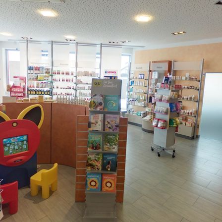 Ein Apothekeninnenraum zeigt Bücher und Produkte. Regale mit Artikeln säumen die Wände. Bunte Stühle und ein Tisch im Vordergrund. Eine helle Decke mit Lichtern.