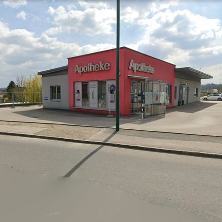 Eine Apotheke befindet sich an einer Straße mit einem Parkplatz. Die Apotheke hat eine rote Fassade mit großen Fenstern und Schildern, die 'Apotheke' lesen. Ein Straßenlicht ist auf der linken Seite sichtbar. In der Ferne befindet sich ein weißes Auto und ein Baum.