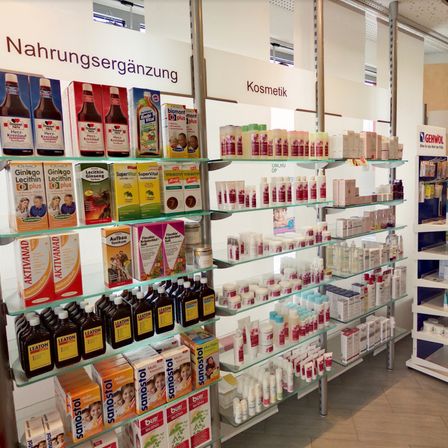 Eine Apotheke mit Regalen, die verschiedene Produkte wie Vitamine, Nahrungsergänzungsmittel und Kosmetikartikel ausstellt. Die Regale haben einen weißen Hintergrund mit Etiketten.