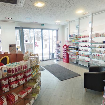 Innenansicht eines Geschäfts mit mehreren Regalen, gefüllt mit Produkten, einer Glastür und einem großen Fenster. Ein schwarzer Teppich liegt vor einem schwarzen Stuhl.