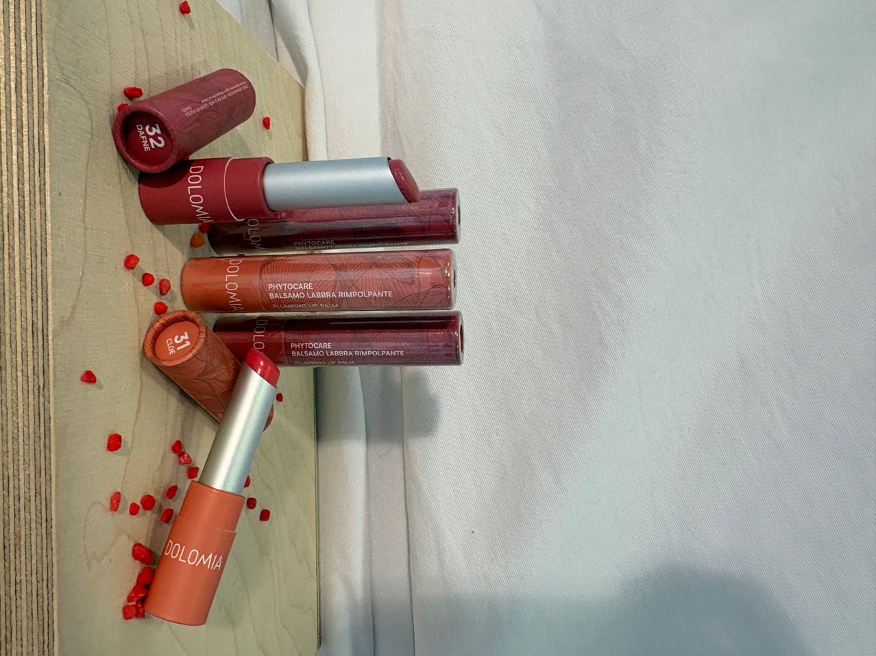 Sechs rote Lippenstifte von Dolomia mit roten Herzen darum, gestapelt auf einer hölzernen Oberfläche mit einem weißen Tuch im Hintergrund.