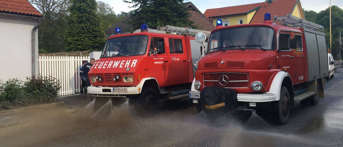 Zwei rote Feuerwehrfahrzeuge sprühen Wasser auf die Straße. Eines hat die Aufschrift 'FEUERWEHR' an der Seite. Hinter den Fahrzeugen stehen Bäume und Häuser.
