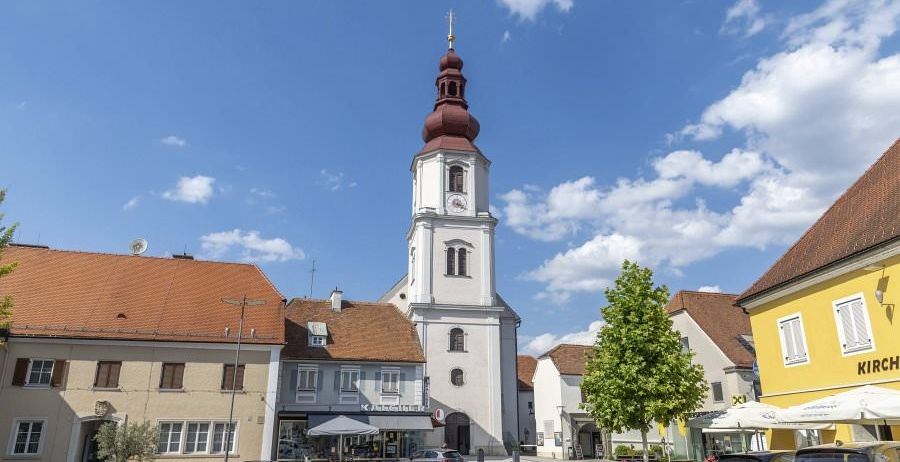 Ein Stadtplatz mit einem weißen Kirchturm und einem roten Dach. Eine Uhr befindet sich auf dem Turm und zwei Autos sind davor geparkt.