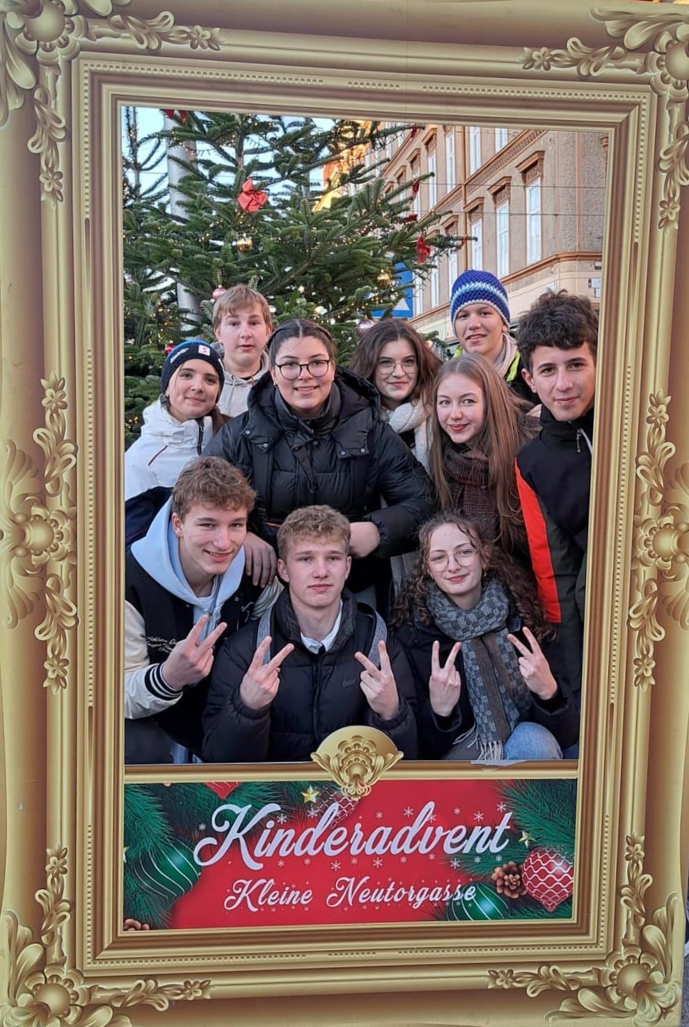 Eine Gruppe junger Menschen posiert für ein Foto vor einem geschmückten Weihnachtsbaum mit einem goldenen Rahmen. Sie alle lächeln und machen Friedenszeichen.