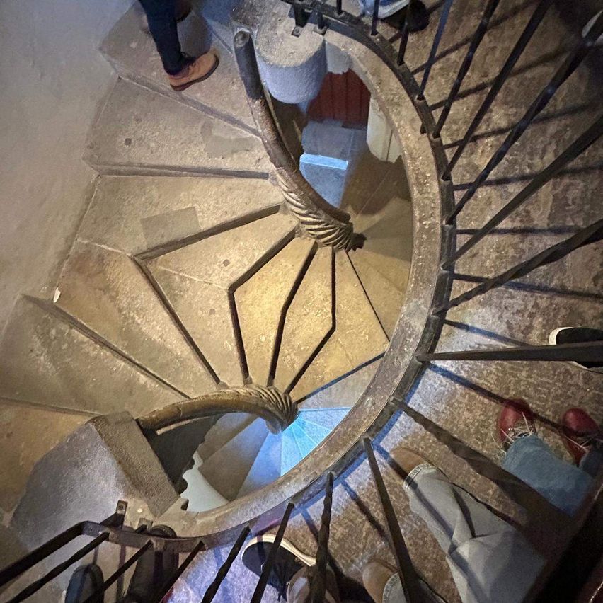 Eine Wendeltreppe mit Steinstufen und Metallgeländern, von unten betrachtet. Die Treppe hat ein kreisförmiges Design mit blauer Beleuchtung am Boden.