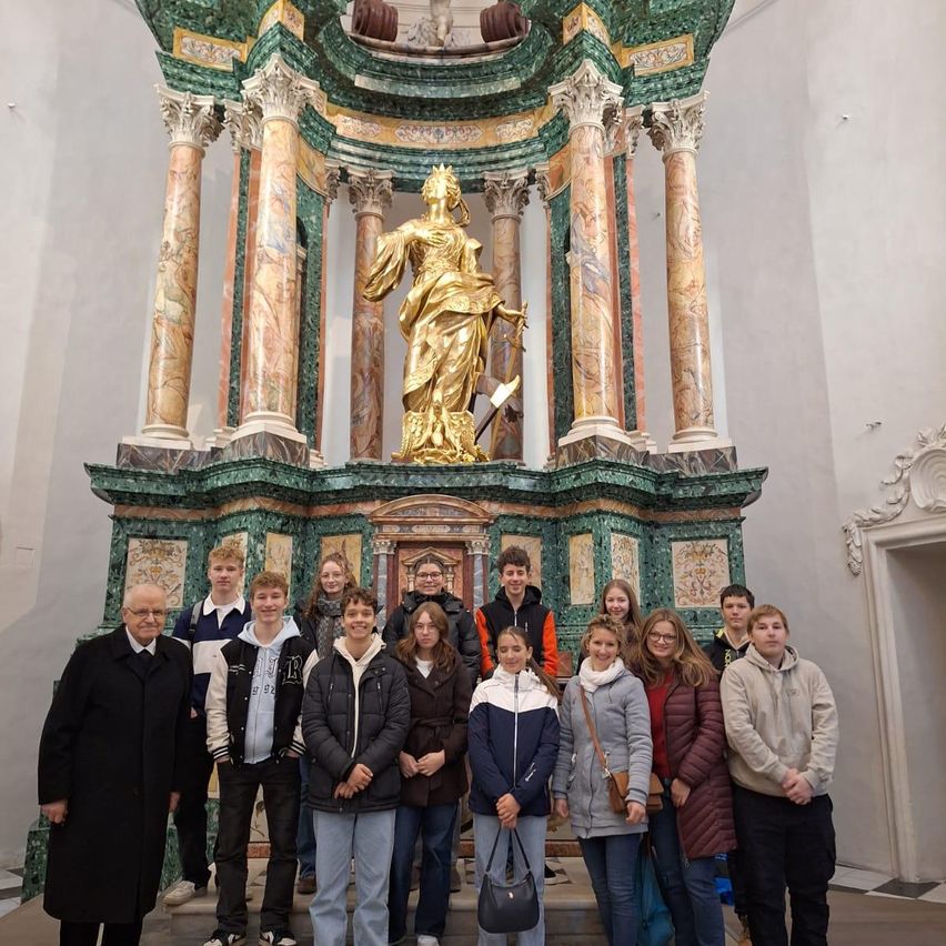 Eine Gruppe von Schülern und einem Lehrer steht vor einer goldenen Statue in einem kunstvollen Raum mit Marmorsäulen.