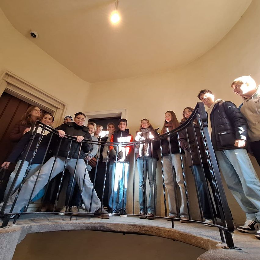 Eine Gruppe von Schülern steht in einem runden Raum und blickt durch eine Wendeltreppe nach unten. Sie tragen Jacken und einige tragen eine Brille. Eine Kamera ist an der Decke montiert.