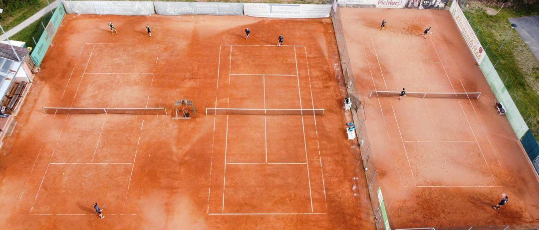 Eine Luftaufnahme eines Tennisplatzes mit Spielern in verschiedenen Positionen. Einige stehen, andere schlagen den Ball. Der Platz ist von einem Zaun umgeben.