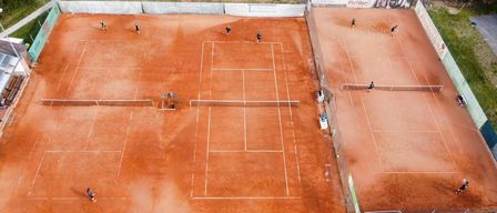 Ein Luftbild eines Tennisplatzes mit Spielern in Aktion. Mehrere Courts sind sichtbar, jeweils mit zwei Spielern. Die Courts sind von einem Zaun und Werbung umgeben.