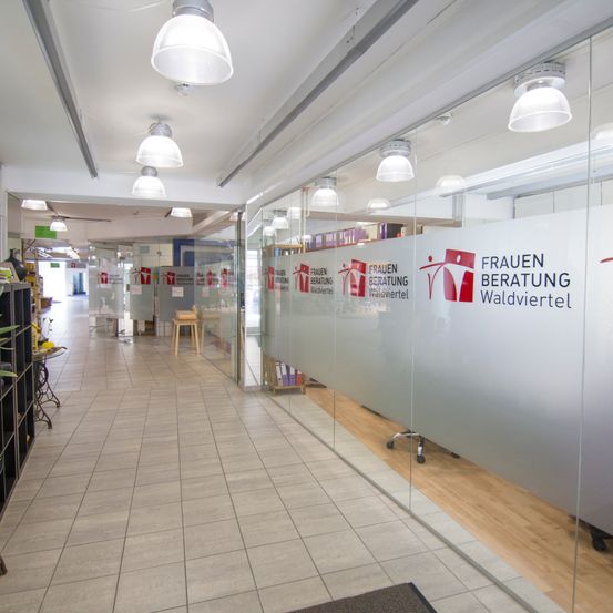 Ein leerer Flur mit weißen Fliesen, Glaswänden und weißen Decken. Die Glaswände haben ein Logo und den Text 'Frauenberatung Waldviertel'. Im Hintergrund befinden sich Tische, Regale und ein Hocker.