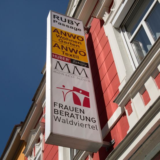 Ein Schild für Ruby Passage, das Damenbekleidung, Heimtextilien und Gärten anbietet. Es ist an einem roten Gebäude mit weißen Abschlüssen befestigt.