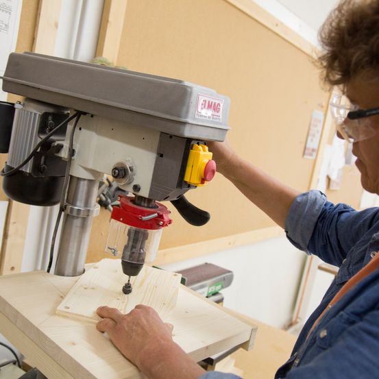 Eine Frau mit Schutzbrille benutzt eine Bohrmaschine, um in ein Stück Holz zu bohren. Die Bohrmaschine ist mit "JMAG" beschriftet und auf einem Holzarbeitstisch montiert.