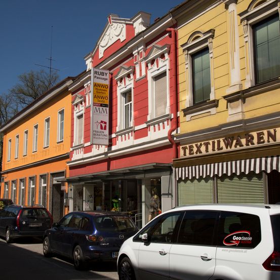 Ein Blick auf die Straße mit geparkten Autos vor Gebäuden, von denen eines ein Schild mit der Aufschrift 'RUBY Passage' hat. Das Gebäude hat eine rot-weiße Fassade.