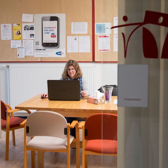 Eine erwachsene Frau sitzt vor einem Laptop auf einem Schreibtisch in einem Büro. Papiere und ein Schild hängen an der Wand hinter ihr.