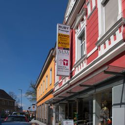 Ein Laden mit roter Wand und Vordach zeigt ein Schild für Ruby Passage und zwei Gärten. Ein Mannequin mit einem Schal ist durch das Fenster sichtbar.