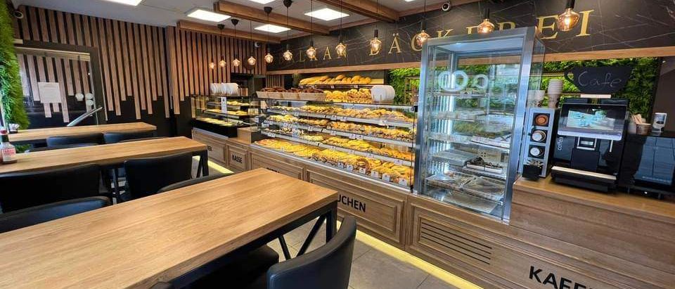 Eine Bäckerei mit Holztischen und Stühlen. Die Vitrine hat verschiedene Gebäcke. Der Laden hat eine Holztheke und eine Kaffeemaschine.