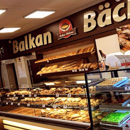 Ein Bäckereiladen zeigt verschiedene Brote, Gebäck und Kuchen in Glasvitrinen. Das Schild heißt 'Balkan' und hat ein Logo. Der Laden hat eine Uhr an der Wand.