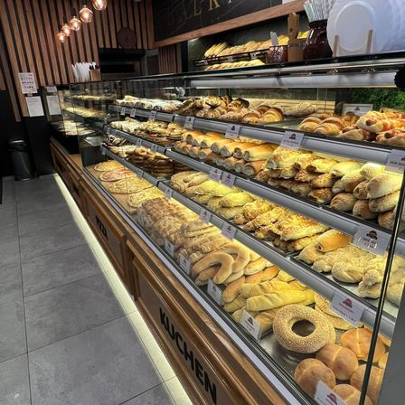 Eine Bäckerei-Vitrine mit verschiedenen Gebäcksorten, darunter Brötchen, Croissants und Bagels. Die Vitrine ist von oben mit warmer Beleuchtung beleuchtet.