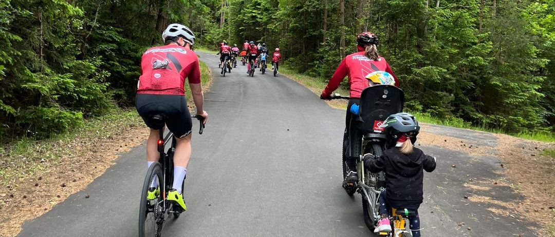 Eine Gruppe von Radfahrern fährt auf einer Straße mit einem Kind in einem Fahrradsitz vorne. Bäume säumen die Straße.