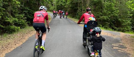Eine Gruppe von Radfahrern fährt auf einer Straße mit einem Kind in einem Fahrradsitz vorne. Bäume säumen die Straße.
