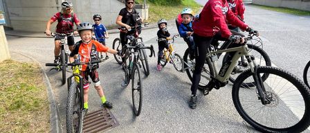 Eine Gruppe von Radfahrern, darunter Erwachsene und Kinder, fährt auf einer Straße mit ihren Fahrrädern. Sie tragen alle Helme und fahren verschiedene Arten von Fahrrädern.