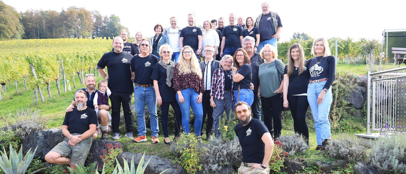 Eine Gruppe von Menschen, in schwarzen T-Shirts gekleidet, steht in einem Weinberg, einige sitzen auf Felsen. Topfpflanzen sind um sie herum.