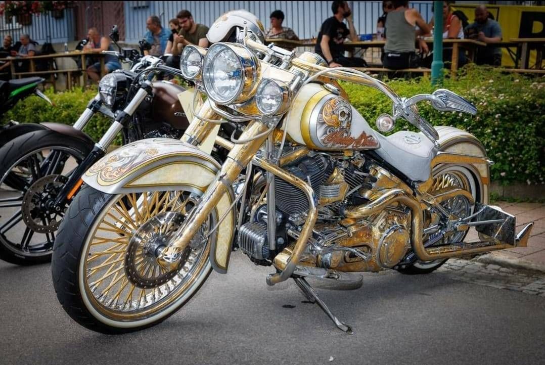 Ein goldenes und weißes Harley Davidson Motorrad geparkt auf der Straße mit Leuten, die um Tische sitzen hinten.