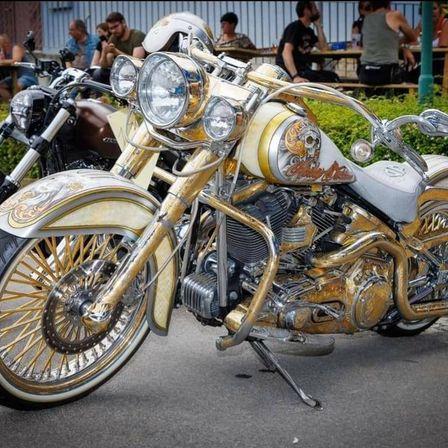 Ein goldenes und weißes Harley Davidson Motorrad geparkt auf der Straße mit Leuten, die um Tische sitzen hinten.
