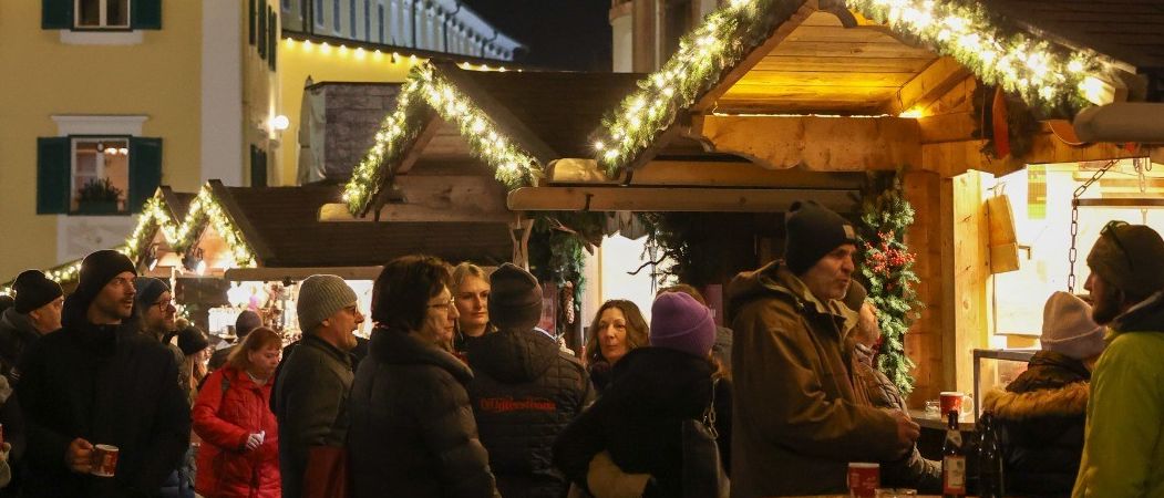 Menschen in Winterkleidung versammeln sich nachts auf einem festlichen Markt, umgeben von Ständen mit Weihnachtslichtern und Dekorationen. Ein Mann steht in der Nähe eines Tisches mit Getränken, andere halten Tassen. Gebäude mit architektonischen Details sind im Hintergrund zu sehen.