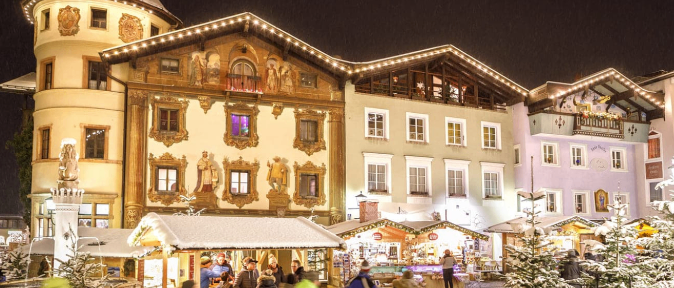 Bei Nacht ist ein Weihnachtsmarkt in einem Stadtplatz belebt. Menschen gehen an geschmückten Ständen und Gebäuden mit kunstvollen Fassaden vorbei. Schnee bedeckt den Boden und die Bäume.