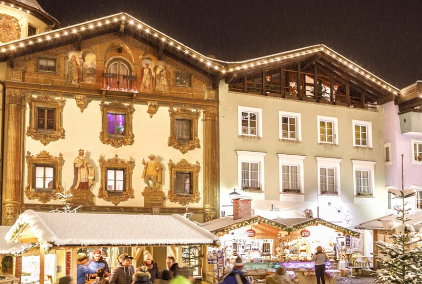 Bei Nacht ist ein Weihnachtsmarkt in einem Stadtplatz belebt. Menschen gehen an geschmückten Ständen und Gebäuden mit kunstvollen Fassaden vorbei. Schnee bedeckt den Boden und die Bäume.