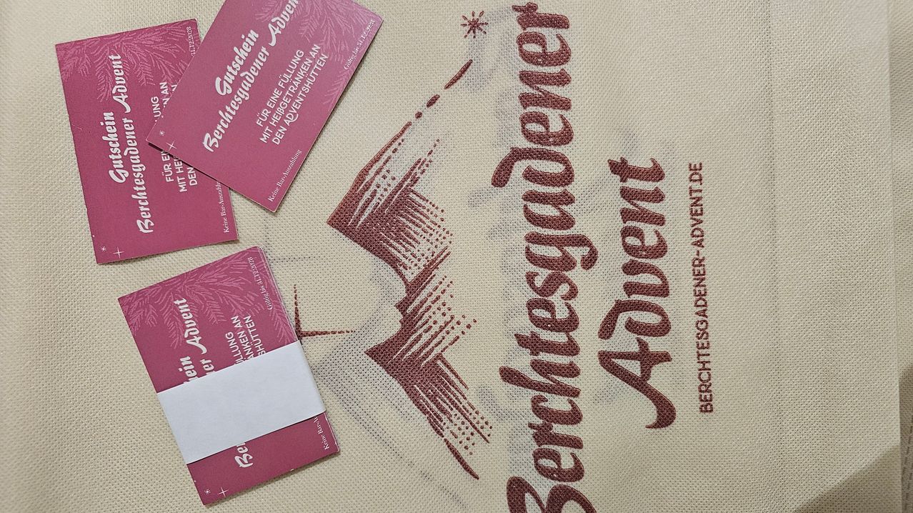 A close-up view of a beige fabric featuring the word Abenteuer in red cursive font. Beside it, there are two small, pink-colored booklets with white text and a white logo on the bottom left.