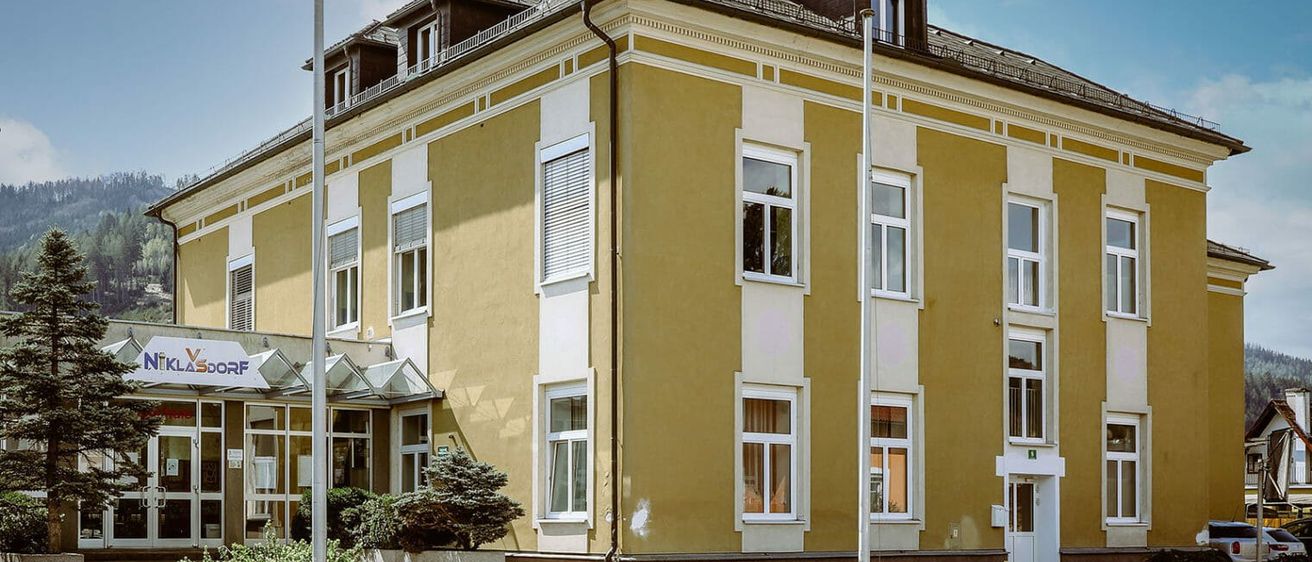 Ein gelbes Gebäude mit mehreren Fenstern und einem kleinen Vordach, mit Büschen und einem Metallzaun davor.