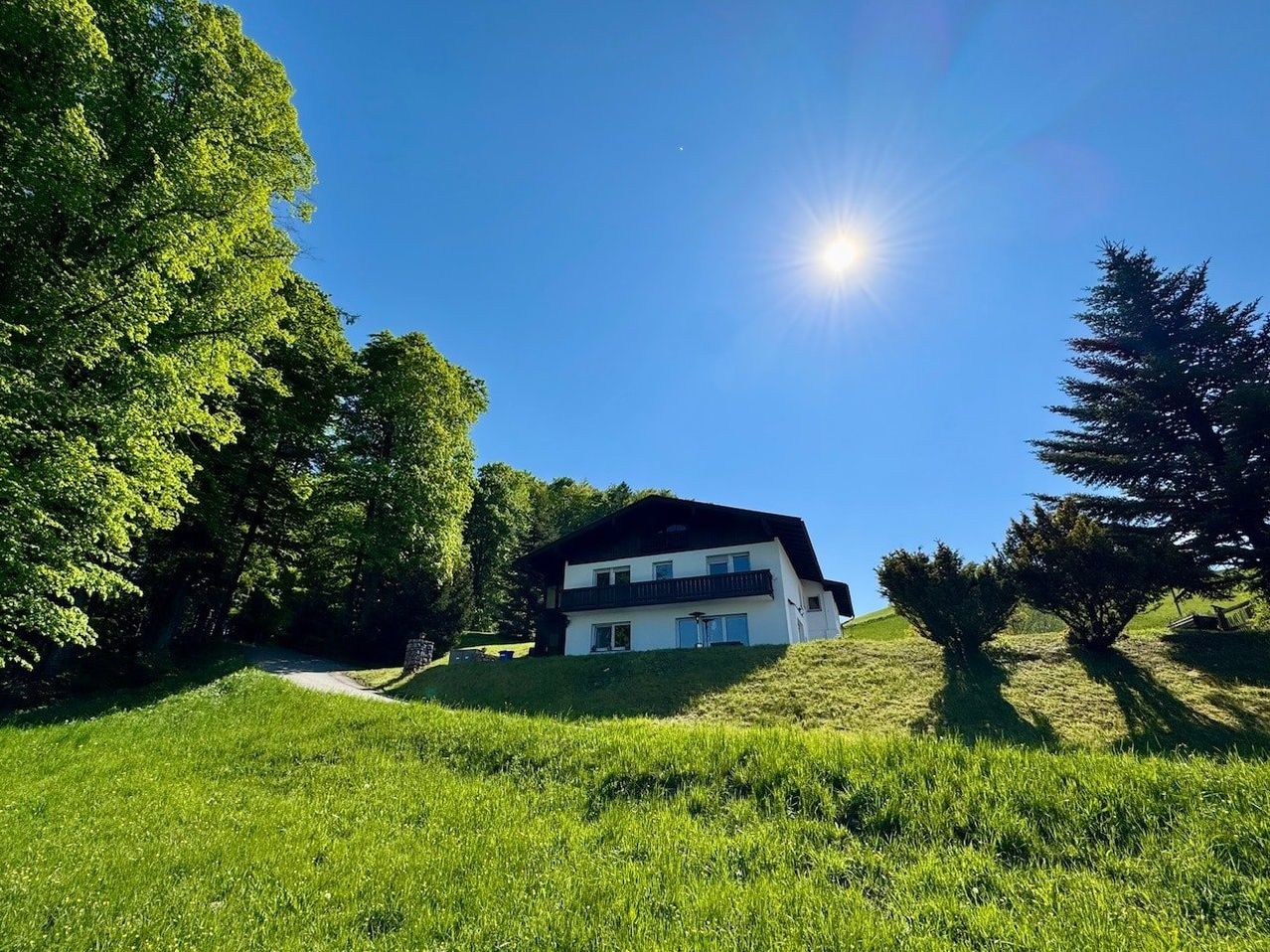 An einem sonnigen Tag zeigt ein weißes Haus mit schwarzem Dach, eingebettet auf einem Grashügel, umgeben von Bäumen.