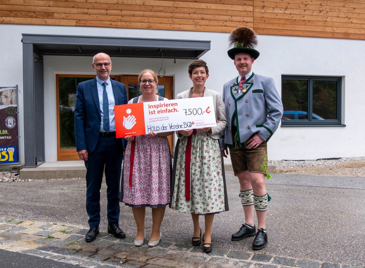 Vier Personen stehen vor einem Gebäude und halten ein großes Schild mit der Aufschrift 'Inspireren ist einfach. 1.7500,-'. Zwei Frauen tragen traditionelle Kleider und ein Mann ein traditionelles Outfit. Hinter ihnen steht ein Mann im Anzug.