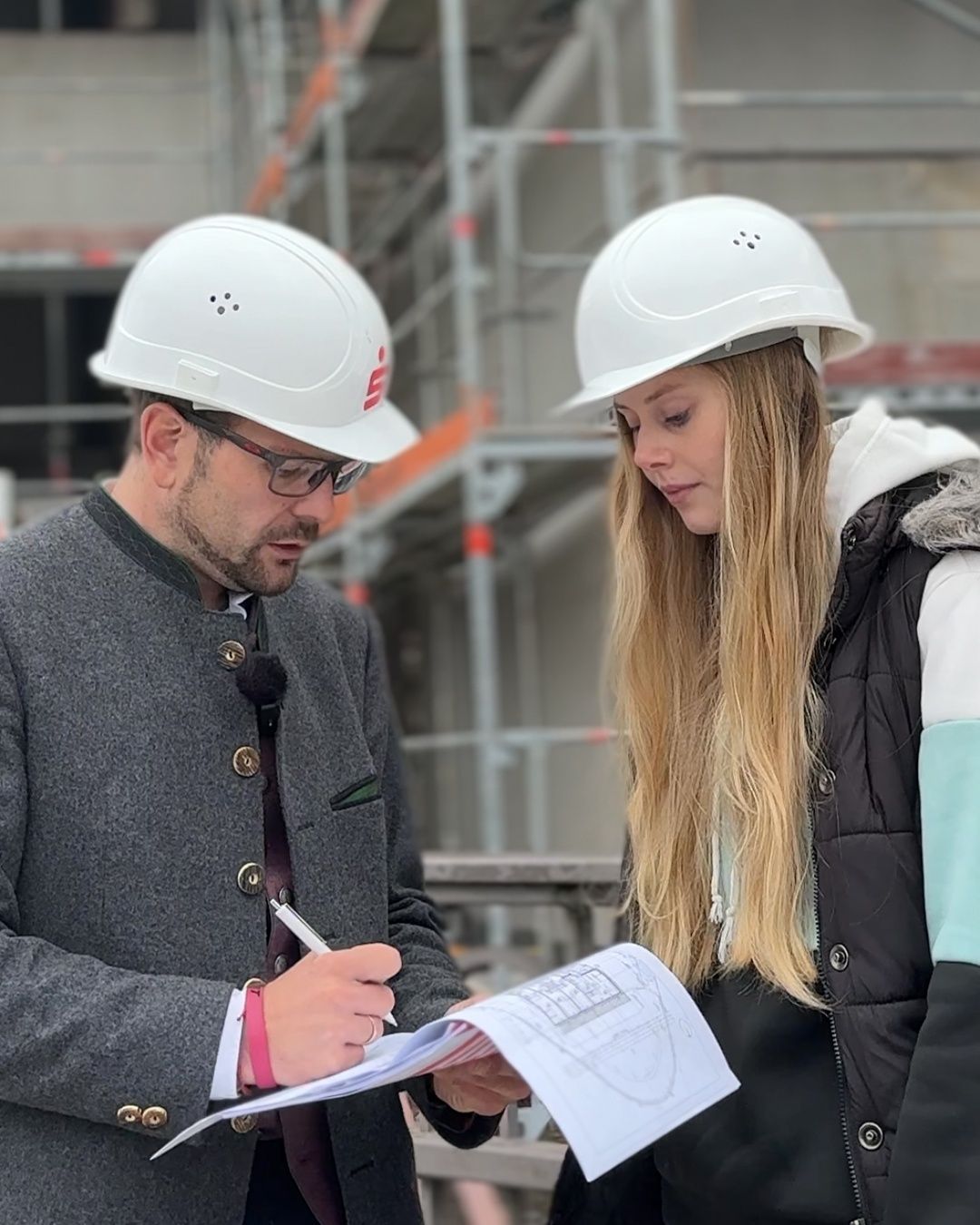 Zwei Personen in weißen Schutzhelmen überprüfen Baupläne. Der Mann schreibt, während die Frau aufmerksam zusieht. Sie stehen auf einer Baustelle.